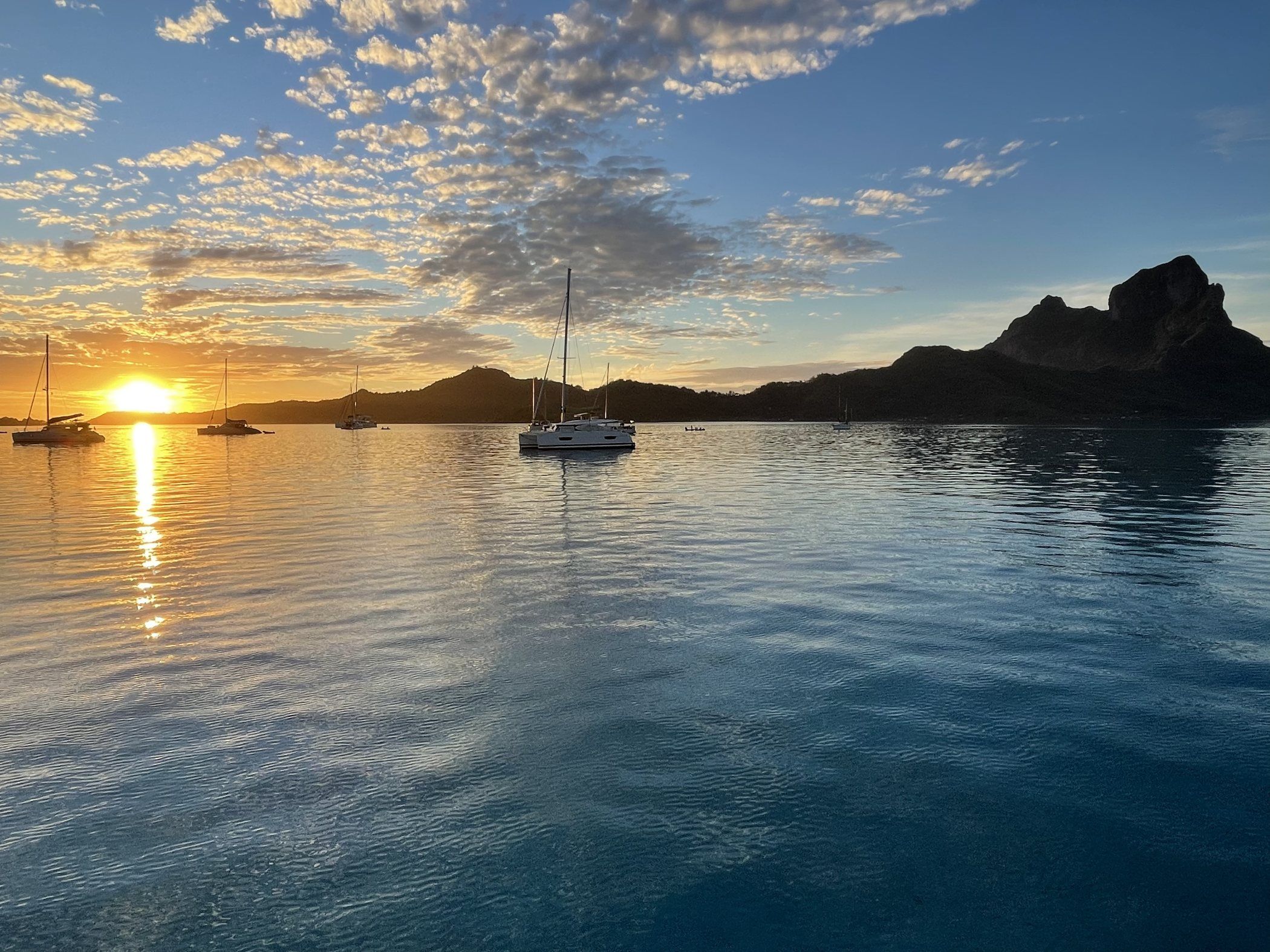 A catamaran at anchor on a still lagoon at sunset, Bora Bora's mountain silhouette in the distance.