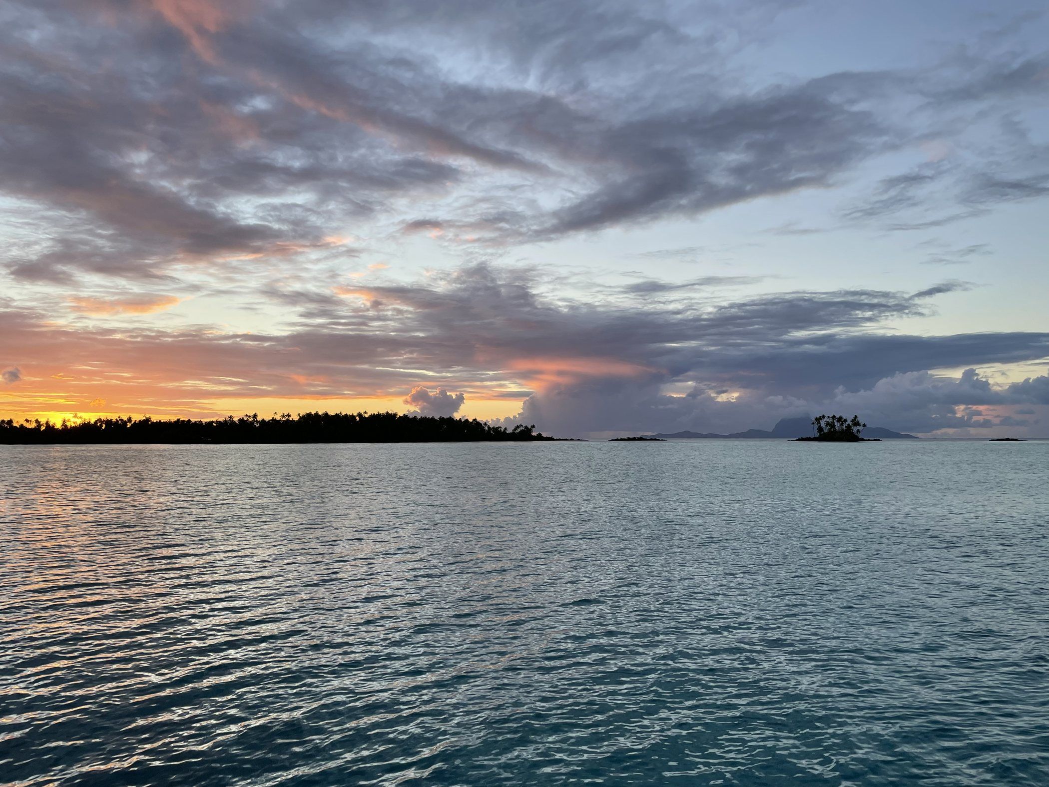 A still Polynesian lagoon at dusk, low palm-lined islands silhouetted under a violet and amber sky.