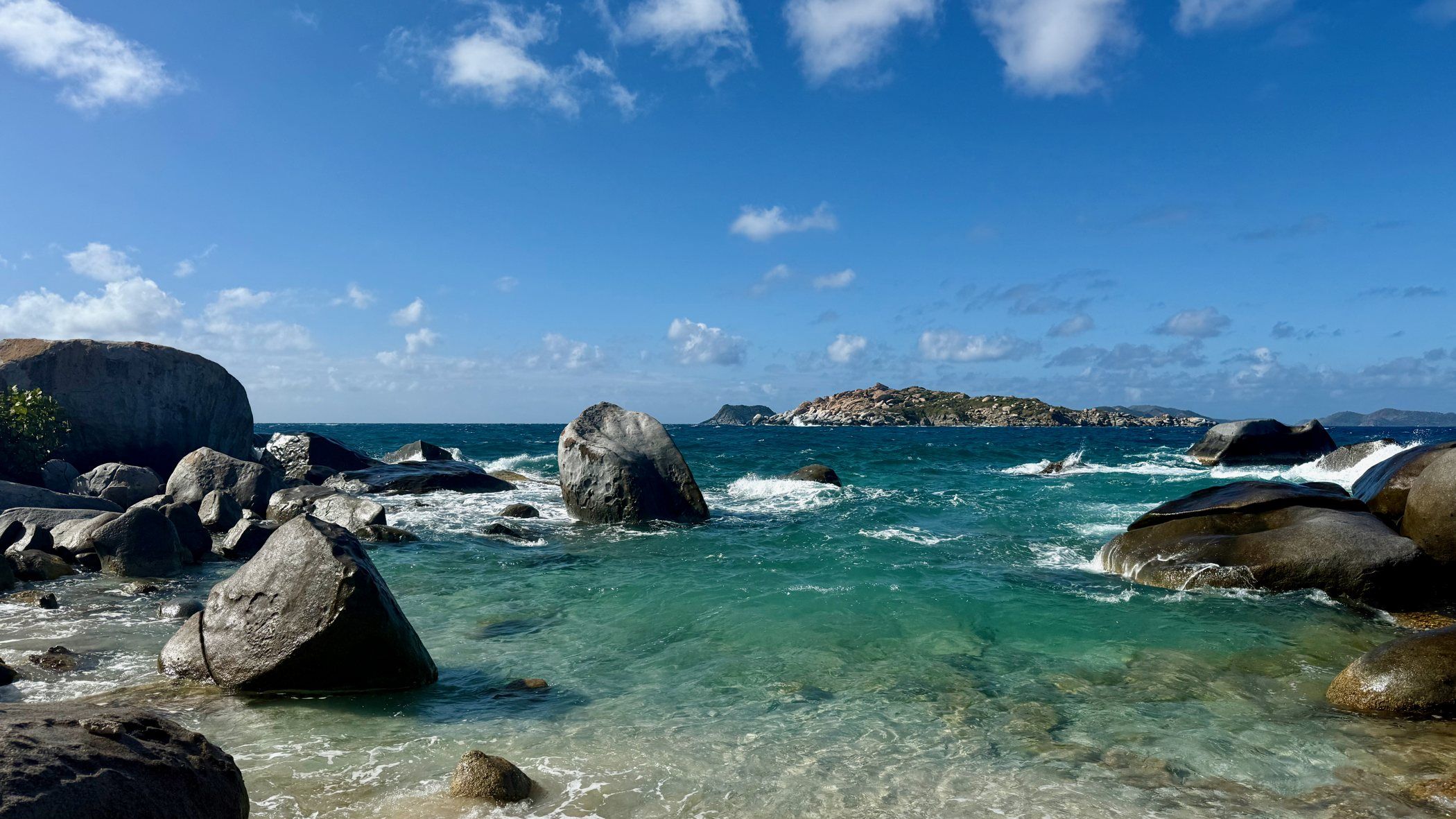 Rounded granite boulders standing in shallow turquoise surf at The Baths, a low island across the channel under a bright sky.